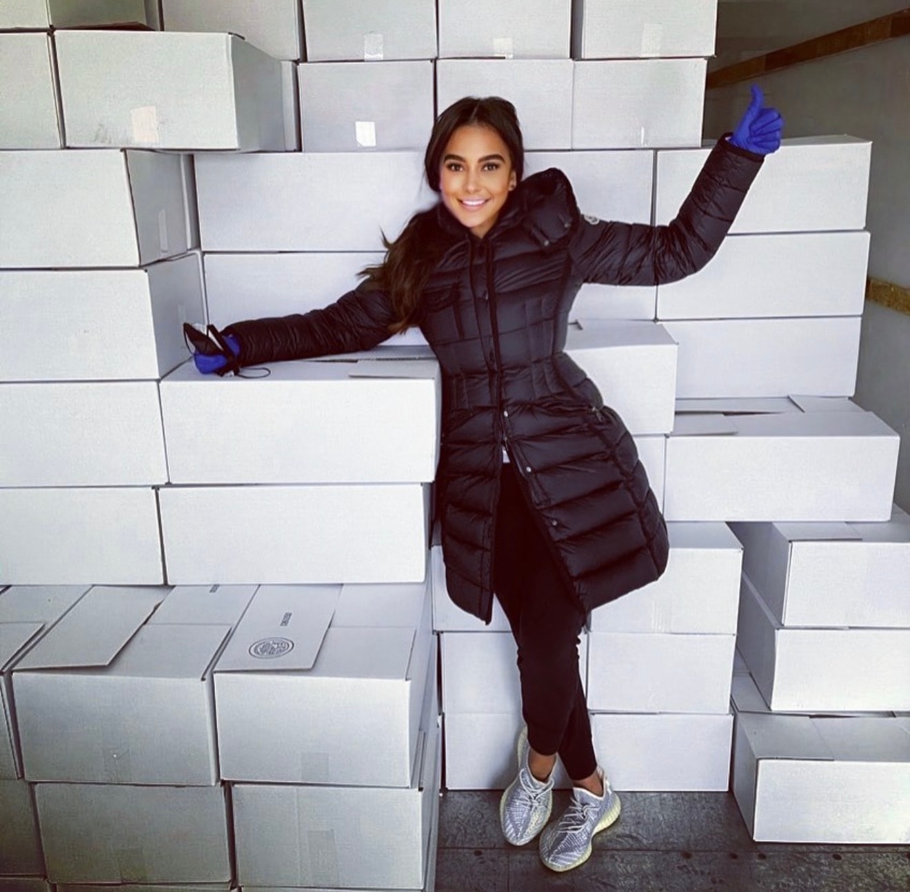 Talia Ramos at a community service event among stacked white boxes, smiling with raised hand